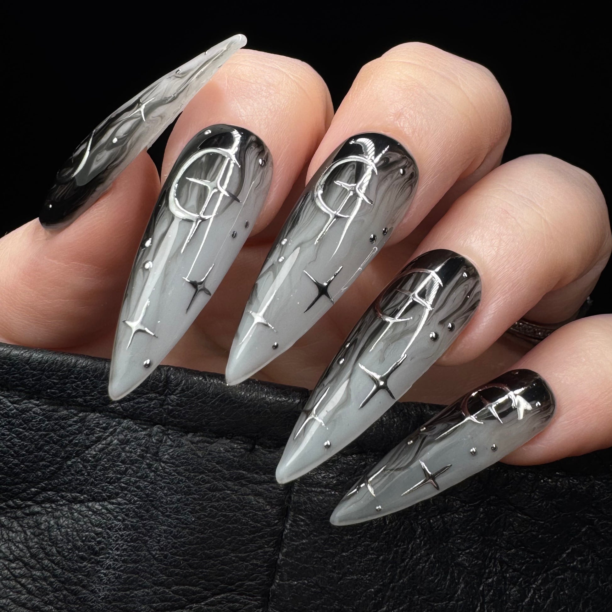 A close-up of a hand with long stiletto-shaped nails featuring a black-to-white gradient design with silver metallic line art and small metallic dots, resting on a black textured surface.