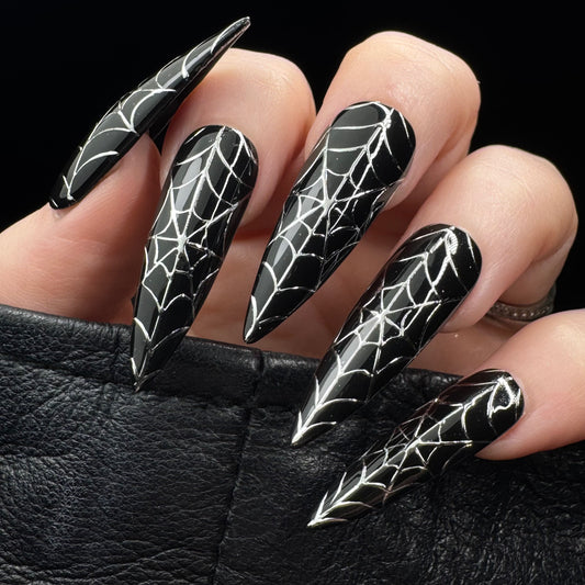 A close-up of a hand with long stiletto-shaped nails painted black and decorated with silver spiderweb patterns, resting against a black textured surface.